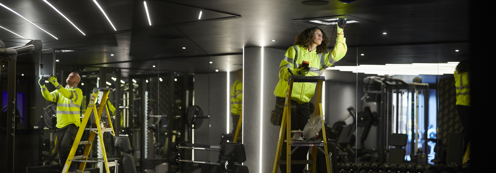 Two technicians on ladders inspecting ceiling-mounted building equipment